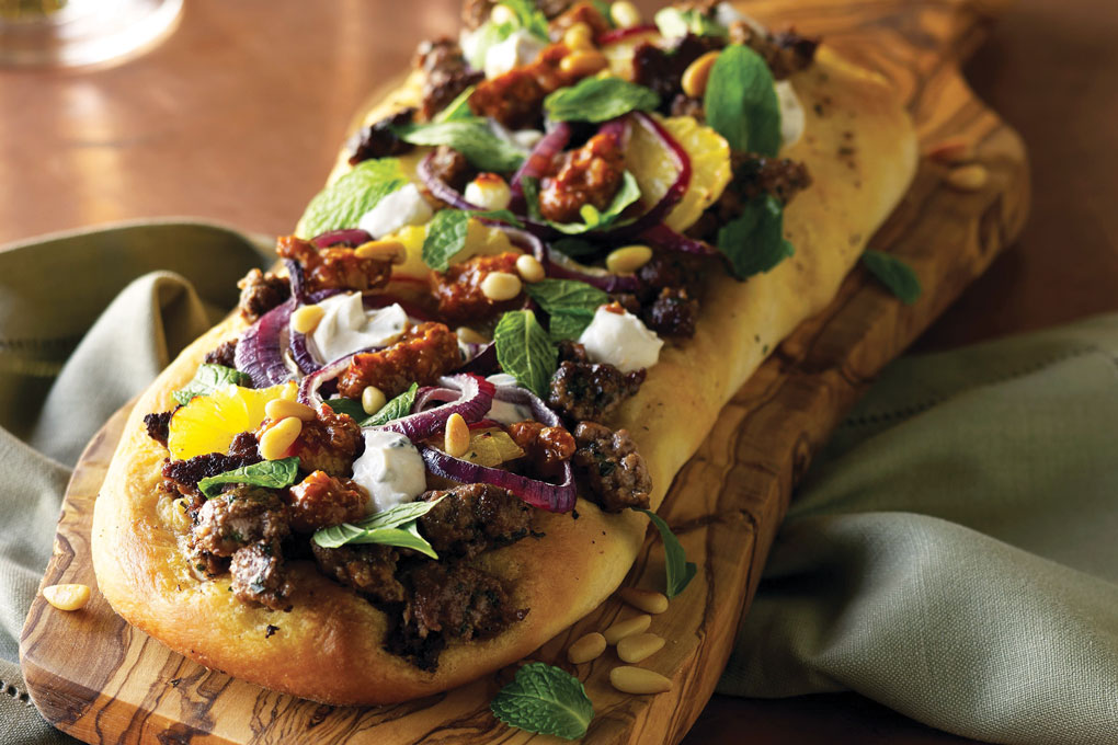 A drizzle of tahini yogurt and harissa completes this Lamb Merguez Flatbread, where ground lamb and Middle Eastern spices top a toasted flatbread. Fresh mint and pine nuts finish the dish.