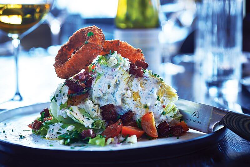 The Wedge becomes a backdrop for craveable inclusions at Maple & Ash. Crisp iceberg holds up spicy onion rings and crispy glazed bacon, along with marinated heirloom cherry tomatoes.