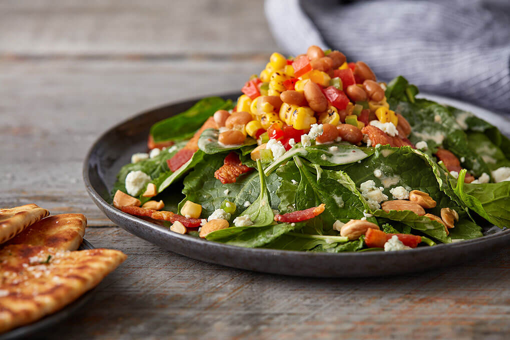 This spinach and kale salad gets a flavor upgrade with lardons, bleu goat cheese and Marcona almonds, all tossed with a warm bean and bacon dressing and topped with baked bean “cowboy caviar.”