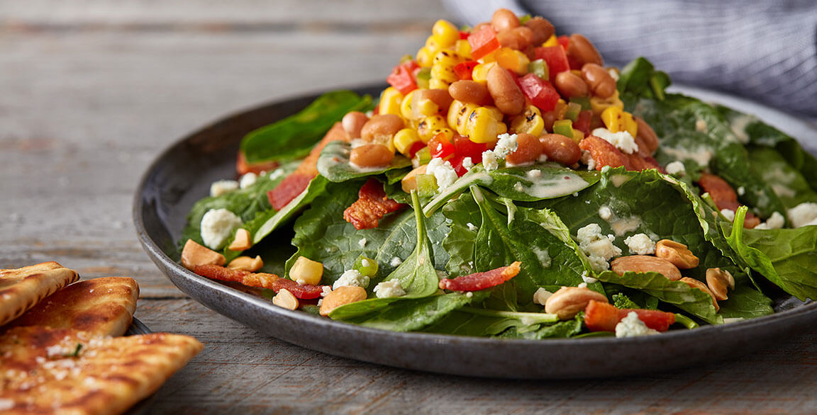 This spinach and kale salad gets a flavor upgrade with lardons, bleu goat cheese and Marcona almonds, all tossed with a warm bean and bacon dressing and topped with baked bean “cowboy caviar.”