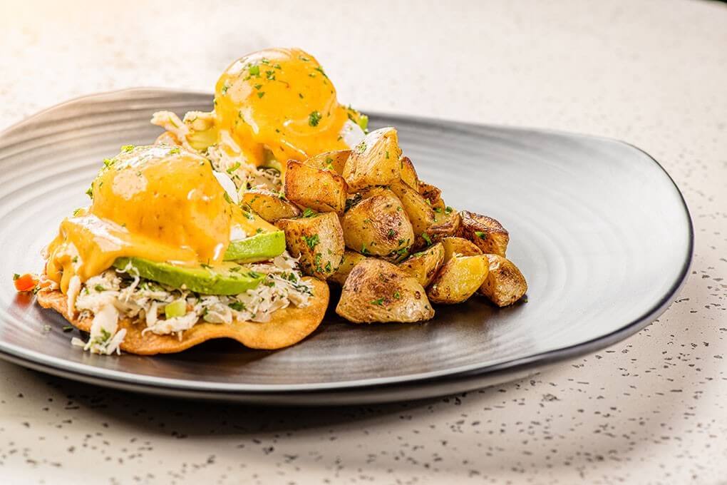 Crispy-fried white corn tortillas topped with refried beans, smashed avocado, crab salad and a soft-poached egg, finished with chipotle hollandaise, served with papas rancheras