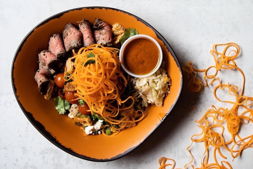 Spice-rubbed steak with spinach, roasted potatoes and peppers, corn, feta, mango chutney, spiralized carrots, coleslaw and green onions, served with a mango-chipotle sauce
