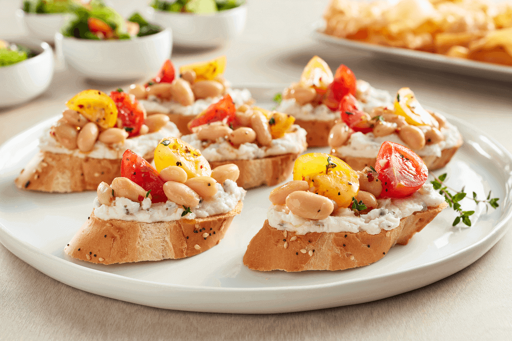 : Rich and creamy dairy-free crostini with Boursin® Dairy-Free Cheese Spread Alternative, Garlic & Herbs, topped with cannellini beans, minced shallots, cherry tomatoes and fresh thyme.